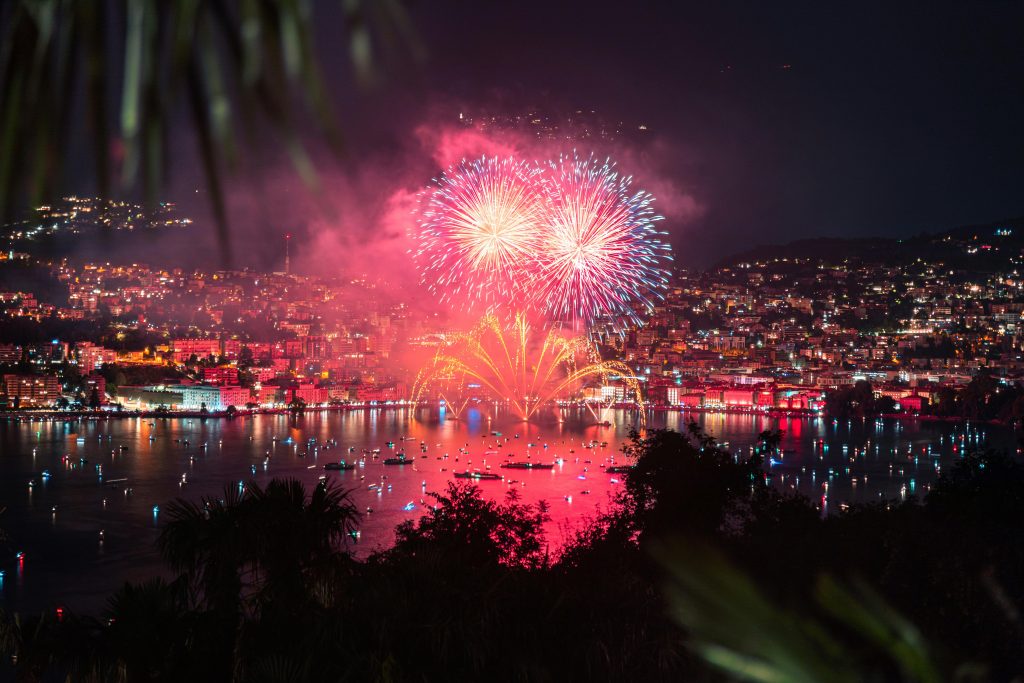 Vibrant fireworks lighting up the night sky over a coastal city and harbor, with reflections shimmering on the water.
