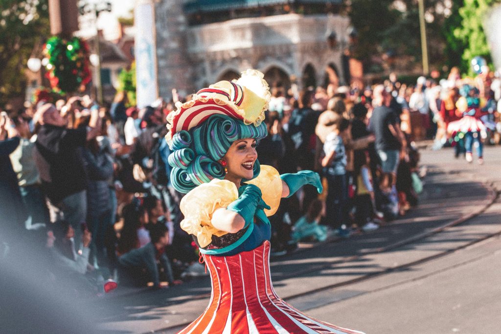 Performer in a colorful costume with a teal wig and candy-striped dress dancing in a Disney parade with a cheering crowd in the background.